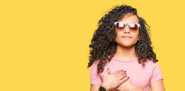 Kıvırcık saçlı pembe ile gülümseyen güneş gözlüğü takmış genç güzel kadın kapalı gözler ve yüzünde minnettar jest ile göğüs üzerinde eller. Sağlık kavramı.