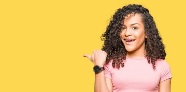 Kıvırcık saçlı pembe tişört arıyorum ve başparmak yanında yukarıyı mutlu yüz ile gülümseyen giyen genç güzel kadın.