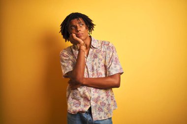 Tatil dreadlocks ile Afro adam izole sarı arka plan üzerine el çene hakkında soru, dalgın ifade düşünme ile çiçek gömlek giyiyor. Düşünceli bir yüz ile gülümsüyor. Şüphe kavramı.