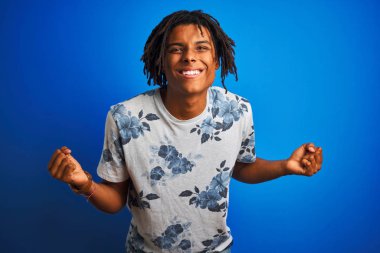 Afro Amerikan adam dreadlocks ile izole mavi arka plan üzerinde yaz çiçek t-shirt giyiyor şaşırttı ve silah yükseltilmiş ve açık gözleri ile başarı için hayran kutluyor. Kazanan kavramı.