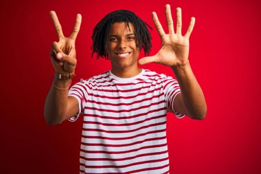 Çizgili t-shirt giyen dreadlocks giyen genç afro adam izole kırmızı arka plan üzerinde duran gösteren ve emin ve mutlu gülümserken parmakları sayısı yedi ile işaret.