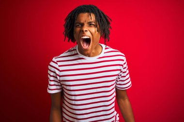 Çizgili t-shirt giyen dreadlocks giyen genç afro adam izole kırmızı arka plan üzerinde duran kızgın ve deli çığlık sinirli ve öfkeli, öfke ile bağırarak. Öfke ve agresif kavram.