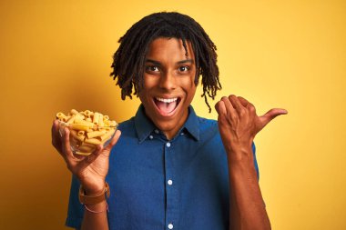 Afro Amerikan adam dreadlocks tutan makarna makarna üzerinde izole sarı arka plan işaret ve başparmak ile yan kadar mutlu yüz gülümseyen ile gösteren
