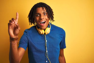 Kıvırcık sarı arka plan üzerinde kulaklık giyen dreadlocks ile Afro Amerikan adam başarılı bir fikir parmak yukarı işaret. Çıkıldı ve mutlu. Bir numara.