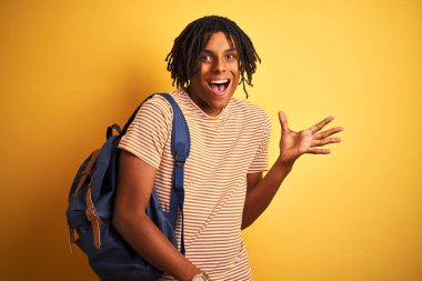 Izole sarı arka plan üzerinde sırt çantası giyen dreadlocks ile Afro Amerikan öğrenci adam çok mutlu ve heyecanlı, kazanan ifade büyük bir gülümseme ile çığlık zafer kutluyor ve ellerini kaldırdı