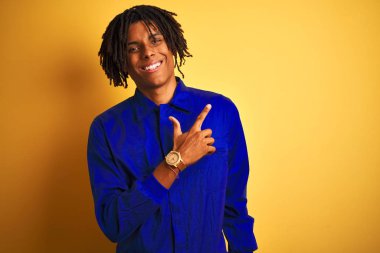 Yüz üzerinde mutlu ve doğal ifade ile yan kadar el ve parmak ile işaret yüz bir gülümseme ile izole sarı arka plan üzerinde mekanik üniforma giyen dreadlocks ile Afro işçi adam
