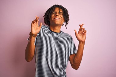 Zencili pembe arka plan üzerinde ayakta lacivert çizgili t-shirt dikilmiş dreadlocks ile Afro adam umut ve gözler ile gülümseyen geçti parmak kapalı. Şans ve batıl fikir.