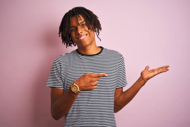 Siyah izole pembe arka plan üzerinde ayakta lacivert çizgili t-shirt giymiş dreadlocks ile Afro adam hayran ve parmak ile işaret ederken kamera gülümseyen.