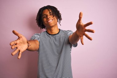 Mor arka plan üzerinde ayakta lacivert çizgili t-shirt giyen dreadlocks ile Afro adam kucaklamak için açık kollar ile gülümseyen kamera bakıyor. Mutluluğu kucaklayan neşeli ifade.