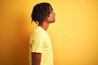 Dreadlocks ile Afro adam izole sarı arka plan üzerinde ayakta rahat t-shirt dikilerek yan arıyor, profil rahat bir gülümseme ile doğal yüz ile poz Relax.