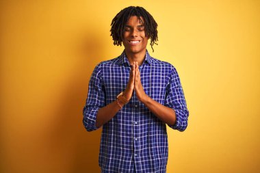Dreadlocks ile Afro adam izole sarı arka plan üzerinde ayakta rahat gömlek dikilerek ellerle birlikte affetmek için güven gülümseyen soran dua.