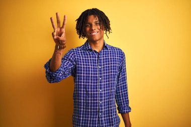 Dreadlocks ile izole sarı arka plan üzerinde duran rahat gömlek ayakta giyen ve parmaklarıyla üç numara ile işaret eden Afro adam emin ve mutlu gülümseyen.