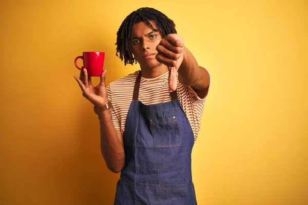 Afro barista man with dreadlocks drinking cup of coffee over isolated yellow background with angry face, negative sign showing dislike with thumbs down, rejection concept