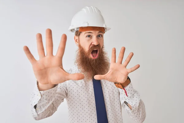 Young redhead irish architect man wearing security helmet over isolated white background afraid and terrified with fear expression stop gesture with hands, shouting in shock. Panic concept.