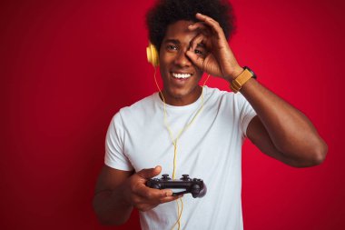 Afro Gamer adam mutlu yüz gülümseyen ile izole kırmızı arka plan üzerinde joystick kulaklıklar kullanarak video oyunu oynarken gözleri parmaklarıyla görünümlü el ile ok işareti yapıyor
