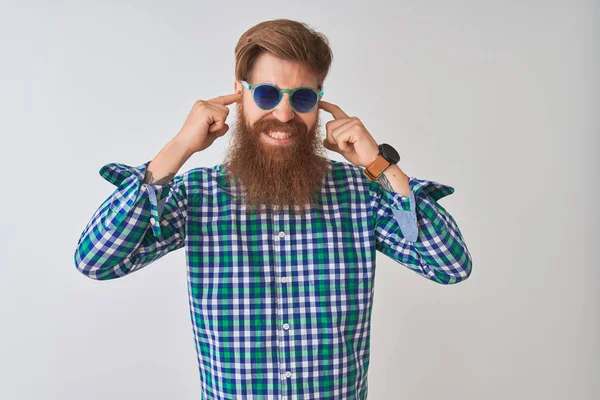 Young redhead irish man wearing casual shirt and sunglasses over isolated white background covering ears with fingers with annoyed expression for the noise of loud music. Deaf concept.