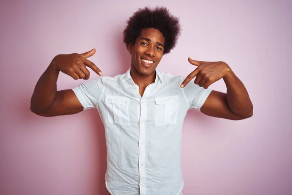 Young american man with afro hair wearing white shirt standing over isolated pink background looking confident with smile on face, pointing oneself with fingers proud and happy.