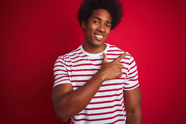年轻的非洲裔美国男子穿着条纹T恤在孤立的红色背景欢快与微笑的脸指向手和手指一边与快乐和自然的表情的脸 — 图库照片
