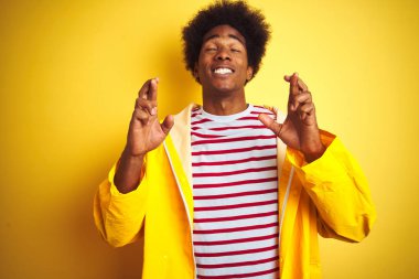 Afro saç ile Afrikalı Amerikan adam izole sarı arka plan üzerinde ayakta yağmur ceket giyiyor umut ve gözler ile gülümseyen geçti parmak kapalı. Şans ve batıl fikir.
