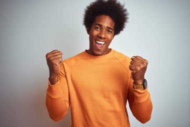 Afro saç ile genç Amerikan adam izole beyaz arka plan üzerinde turuncu kazak giyiyor şaşırttı ve silah yükseltilmiş ve açık gözleri ile başarı için hayran kutluyor. Kazanan kavramı.