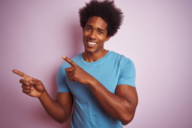 Afro saç ile Afrikalı Amerikan adam izole pembe arka plan üzerinde ayakta mavi t-shirt dikilme gülümseyen ve iki el ve parmak tarafına işaret kamera bakıyor.