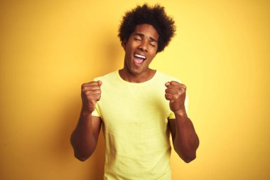 Afro saç ile Afrikalı Amerikan adam izole sarı arka plan üzerinde ayakta t-shirt giyen çok mutlu ve heyecanlı silah kaldırdı, gülümseyen ve başarı için çığlık ile kazanan jest yapıyor. Kutlama konsepti.
