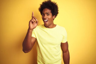 Afro saç ile Afrika Amerikan adam izole sarı arka plan üzerinde ayakta t-shirt giyerek başarılı bir fikir parmak yukarı işaret. Çıkıldı ve mutlu. Bir numara.