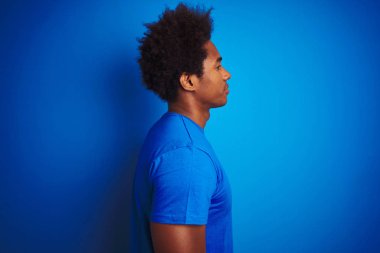 Afro saç ile Afrikalı Amerikan adam izole mavi arka planda duran t-shirt dikili yan bakıyor, rahat bir gülümseme ile doğal yüzü ile profil poz rahatlayın.