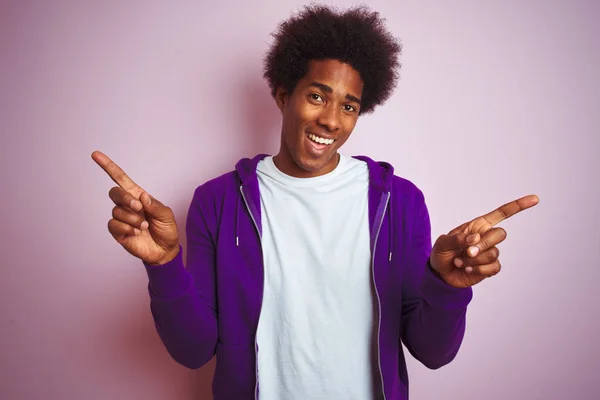 孤立したピンクの背景の上に立つ紫色のスウェットシャツを着た若いアフリカ系アメリカ人男性は 異なる方向に指を向けて自信を持って微笑んでいます 提供情報用のスペースをコピーする — ストック写真
