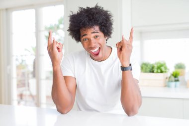 Genç Afrikalı Amerikalı adam rahat beyaz t-shirt evde oturan deli ifade ile eller yukarı kaya sembolü yapıyor bağırıyor. Müzik yıldızı. Ağır müzik konsepti.