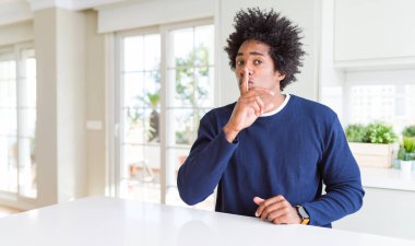 Evde oturan rahat kazak giyen genç Afrikalı Amerikalı adam dudaklarında parmak ile sessiz olmak istiyor. Sessizlik ve gizli konsept.