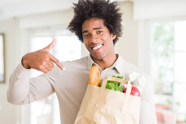 African American man holding groceries bag with fresh vegetables at home with surprise face pointing finger to himself