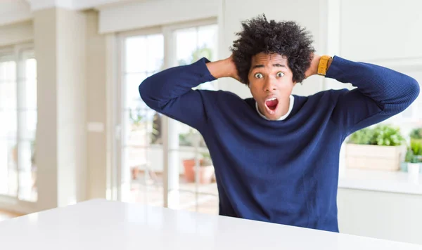 カジュアルなセーターを着た若いアフリカ系アメリカ人男性は 自宅のクレイジーに座って 頭の上に手で怖がって 口を開いてショックを恐れ 驚いた — ストック写真