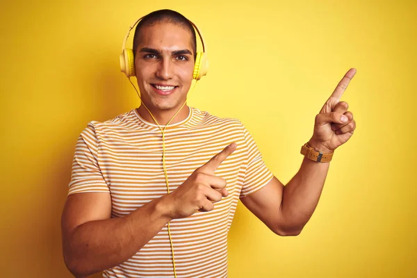 年轻的帅哥使用耳机在黄色孤立的背景微笑 看着相机指着两只手和手指的一侧 — 图库照片