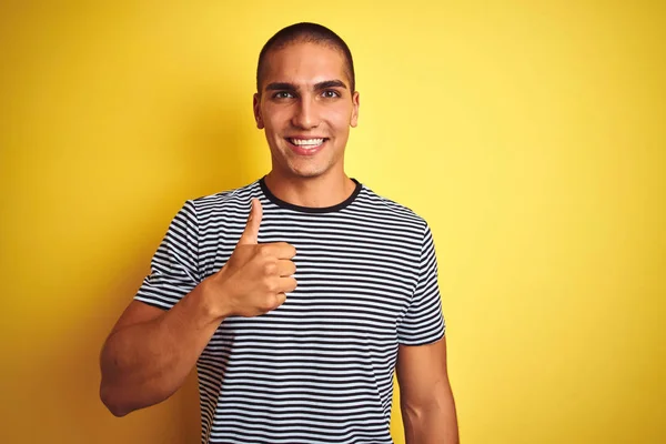 黄色の孤立した背景の上にストライプのTシャツを着た若いハンサムな男は 手で幸せな親指アップジェスチャーを行います 成功を示すカメラを見て式を承認する — ストック写真