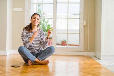 Evde katta oturan genç güzel kadın gülümseyerek ve iki elleri ve parmakları ile tarafına işaret kamera bakıyor.