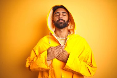 Genç Hint adam kapalı gözler ve yüz minnettar jest ile göğsüne eller ile gülümseyen izole sarı arka plan üzerinde Hood ayakta ile yağmurluk giyiyor. Sağlık konsepti.