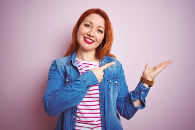 Güzel kızıl saçlı kadın kot gömlek giyiyor ve çizgili t-shirt üzerinde izole pembe arka plan hayran ve kamera için gülümseyen el ile sunmak ve parmak ile işaret.