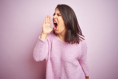 Genç güzel esmer kadın pembe izole arka plan üzerinde bir kazak giyiyor bağırmak ve ağzına el ile yan gürültülü çığlık. İletişim konsepti.