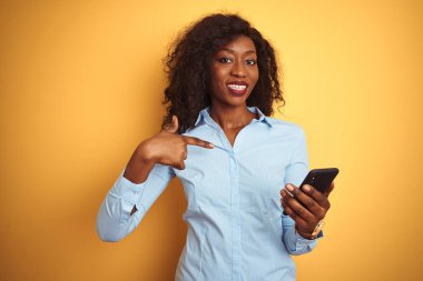 Genç afro-amerikan kadın kendine parmak işaret sürpriz yüzü ile izole sarı arka plan üzerinde duran akıllı telefon kullanarak