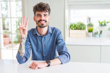 Genç adam rahat gömlek beyaz tablo üzerinde oturan giyen ve parmak numarası dört ile işaret kadar emin ve mutlu gülümseyen.