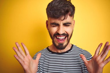 Dövme ile genç adam izole sarı arka plan üzerinde duran çizgili t-shirt dikilmiş silah ile başarı için deli ve çılgın kutluyor ve kapalı gözleri heyecanlı çığlık kaldırdı. Kazanan kavramı