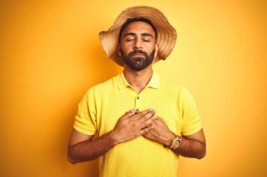 Tatil üzerinde genç Hint adam kapalı gözler ve yüz minnettar jest ile göğsüne eller ile gülümseyen izole sarı arka plan üzerinde ayakta yaz şapka giymiş. Sağlık konsepti.