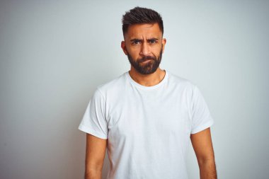 İzole beyaz arka plan üzerinde ayakta t-shirt giyen genç Hint adam şüpheci ve sinir, sorun nedeniyle üzgün çatık. Negatif kişi.