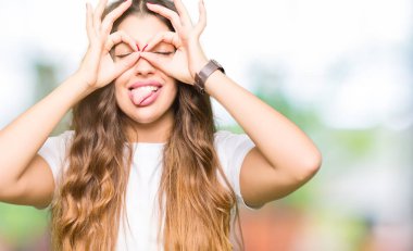 Dilini, parmaklarının arasından baktığını yapışmasını dürbün gibi ok jest yapıyor casual beyaz t-shirt giyen genç güzel kadın. Deli ifade.