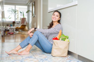 Genç kadın mutfak katında oturan taze bakkallarla dolu bir kağıt çanta yüzünde gülümseme ile yan bakmak, doğal ifade. Güven gülüyor.