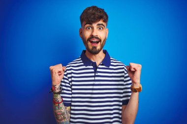 İzole mavi arka plan üzerinde çizgili polo ayakta giyen dövme ile genç adam şaşırttı kutluyor ve başarı için hayran silah kaldırdı ve açık gözler. Kazanan kavramı.