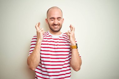 Beyaz izole arka planda işaret parmak üzerinde rahat çizgili kırmızı t-shirt giyen sakal ile genç kel adam umut ve gözler ile gülümseyen geçti kapalı. Şans ve batıl fikir.