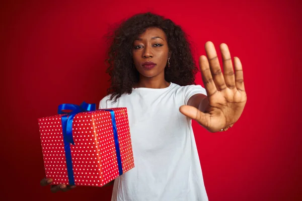 Young african american woman holding birthday gift standing over isolated red background with open hand doing stop sign with serious and confident expression, defense gesture