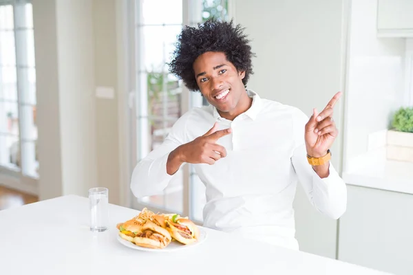 アフリカ系アメリカ人の空腹の男は 昼食のためにハンバーガーを食べて笑顔で 両手と指で側面を指差すカメラを見ている — ストック写真
